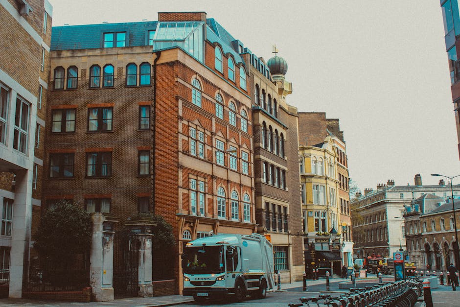 A street scene in an urban area featuring a row of multi-story buildings with diverse architectural styles, including a prominent red-brick building with arched windows, decorative brickwork, and a blue-gray roof with dormer windows. To the right, there is a yellow Victorian-style building with ornate window frames and a decorative top. In the foreground, a white waste disposal truck with blue accents is parked on the cobblestone street, indicating possible involvement in private rubbish removal services. The street is lined with metal bollards and bicycles parked along the sidewalk, with street lamps and a few pedestrians visible further down the road. The sky appears overcast, contributing to diffuse lighting, and the entire scene suggests a busy city centre environment with active waste management operations, including independent collection of rubbish, supporting the context of alternative waste handling outside conventional council services. Waste Disposal Kentish Town’s service is implied through the scene’s focus on rubbish collection activity within this urban setting, consistent with professional waste removal practices.