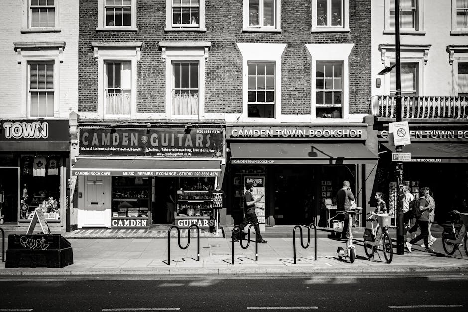A black and white photograph displays the storefronts on a busy street in Kentish Town, with a focus on the Camden Guitar shop on the left, identifiable by its signage advertising musical instruments, repairs, and exchange services. Next to it is the Camden Town Bookshop, shaded by a canopy, with pedestrians walking past and engaging in conversations. Several bicycles are parked along the curb in front of the shops, secured to bike racks. The street scene includes a streetlight and a signpost on the right, with a no-parking sign attached. The multi-story residential building above the shops features a brick exterior, multiple windows with white frames, and some windows have window sills or small decorative ledges. The atmosphere appears overcast, with diffused lighting contributing to the overall neutral tone of the scene, which subtly relates to waste management by illustrating a typical urban shopping area where rubbish collection or on-site clearance services by Waste Disposal Kentish Town might be needed to maintain cleanliness and order in such busy locations.
