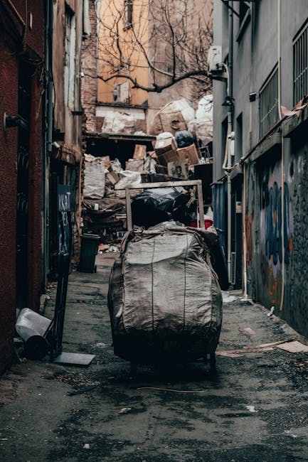 A narrow urban alleyway filled with a large collection of mixed waste and rubbish. In the foreground, there is a prominent grey, weathered wheelie bin with a closed lid, positioned on a rough, dark asphalt surface that shows signs of recent use. Behind the bin, stacked waste bags in black and white are visible along with assorted cardboard boxes and debris, indicating ongoing clearance or disposal activity. The alley is flanked by tall, industrial-style brick and metal buildings, with graffiti visible on a portion of the right-hand wall. Leafless tree branches extend over the scene from the background, and a makeshift wooden barrier or waste collection point can be seen at the far end of the alley, covered with additional stacked refuse. The lighting suggests a cloudy day, casting soft, diffuse illumination over the scene. This setting, associated with private waste handling in an urban environment, reflects the type of site where independent rubbish collection services by Waste Disposal Kentish Town might operate to manage non-domestic waste efficiently and responsibly.