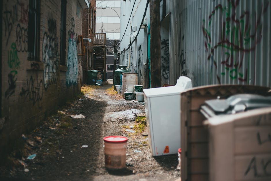 A narrow urban alleyway filled with a large collection of mixed waste and rubbish. In the foreground, there is a prominent grey, weathered wheelie bin with a closed lid, positioned on a rough, dark asphalt surface that shows signs of recent use. Behind the bin, stacked waste bags in black and white are visible along with assorted cardboard boxes and debris, indicating ongoing clearance or disposal activity. The alley is flanked by tall, industrial-style brick and metal buildings, with graffiti visible on a portion of the right-hand wall. Leafless tree branches extend over the scene from the background, and a makeshift wooden barrier or waste collection point can be seen at the far end of the alley, covered with additional stacked refuse. The lighting suggests a cloudy day, casting soft, diffuse illumination over the scene. This setting, associated with private waste handling in an urban environment, reflects the type of site where independent rubbish collection services by Waste Disposal Kentish Town might operate to manage non-domestic waste efficiently and responsibly.