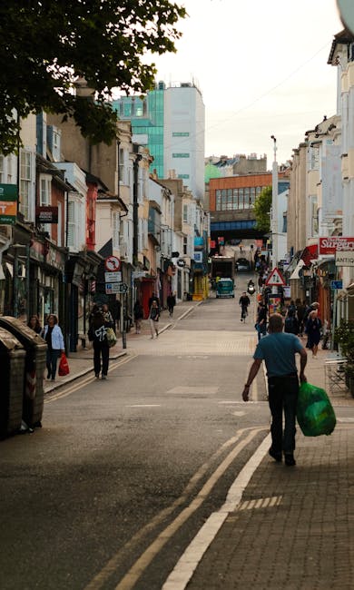 A busy street scene in an urban area with a mix of older and modern multi-story buildings on both sides of the road. The foreground features a man walking away from the camera, carrying a large green bag of waste or recyclable materials. The street is paved with asphalt, with visible road markings and a curb separating the pedestrian walkway. Several pedestrians are walking along the sidewalk, some in groups and others alone, with shopfronts and signs visible on the building facades. In the background, there are taller buildings, including a prominent light blue and white high-rise structure, indicating a commercial or office block. The scene is well-lit with natural daylight, and a large tree with dark green foliage extends into the upper left corner, partially shading the street. The image depicts an area where private or independent waste collection services, such as those offered by Waste Disposal Kentish Town, could be involved in rubbish removal, supporting the local economy and non-council associated waste handling activities.
