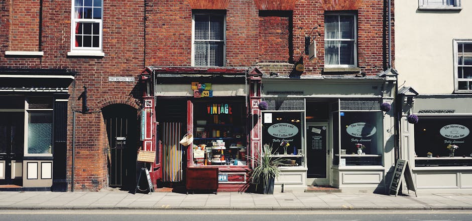 A street scene showing a corner café with a green awning labeled 'CORNER CAFE' situated on the ground floor of a three-story building with a white facade, black timber framing, and red brick accents. The café has large glass windows allowing visibility of the interior, and a few outdoor tables and chairs are placed in front. Adjacent buildings to the left display similar architectural styles with red brickwork and bay windows. The street is paved with asphalt and features road markings including double yellow lines and a pedestrian crossing. A person holding a shopping bag is crossing the street near the café, while vehicles including a red van and a dark-colored car are parked or driving nearby. The scene is illuminated by daylight with shadows cast on the pavement, and utility poles and wires are visible above. This urban environment exemplifies typical residential-commercial mixed-use areas, where private waste collection services, such as those provided by Waste Disposal Kentish Town, are often employed to manage rubbish from local businesses and households, especially along busy town center streets.