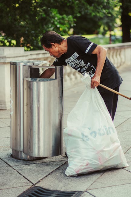 A middle-aged woman with short dark hair is seen disposing of a large white rubbish bag into a modern cylindrical metal bin with a shiny surface on a paved sidewalk. She is wearing a black T-shirt with white text and graphics, and light-colored trousers. She is slightly bent forward, holding a wooden stick in her right hand to assist with lifting or directing the bag. The environment appears to be an outdoor urban area, with a stone balustrade and lush green trees and bushes in the background, indicating a park or public space. Natural daylight illuminates the scene, creating subtle shadows. The woman's actions are typical of private or independent waste disposal, consistent with rubbish removal services provided by Waste Disposal Kentish Town, aimed at maintaining cleanliness and managing waste outside of municipal collection systems.