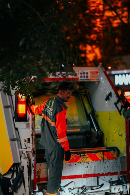 A waste collection operative wearing a high-visibility orange and grey uniform is standing beside a large industrial rubbish collection vehicle during dusk or early evening. The vehicle features a white body with red and yellow reflective markings, and its rear compactor is open, revealing a metal loading area for waste. The background shows silhouetted trees against a sky illuminated by a deep orange sunset, casting a warm glow over the scene. The operative is positioned on a paved surface, possibly a street or parking area, with the vehicle's lights casting a red-orange hue on nearby surfaces. The scene suggests an on-site waste collection operation, consistent with private rubbish removal services, emphasizing the environmental and logistical aspects of alternative waste handling in an urban setting. Waste Disposal Kentish Town is engaged in managing rubbish in an efficient and professional manner, supporting non-council waste disposal methods.