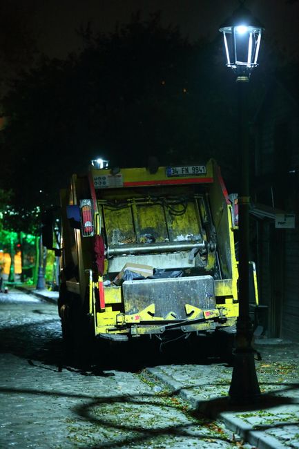 A street scene at night featuring a yellow waste collection vehicle parked on a cobblestone pavement, with a black rear door and mechanical components visible at the back. The vehicle is situated next to a traditional street lamp emitting bright white light, which illuminates part of the surrounding area and casts shadows on the ground. In the background, dark trees and faintly lit buildings are visible, with some green light reflecting off nearby surfaces. The scene suggests an instance of private waste collection or alternative rubbish removal service operating during late hours, with the vehicle positioned in front of a pedestrian walkway or residential area, emphasizing the importance of nighttime rubbish pick-up in urban environments. The overall atmosphere is quiet, with minimal activity or additional objects present, focusing on the waste disposal vehicle and its immediate surroundings, highlighting the operational context of waste management services in Kentish Town.