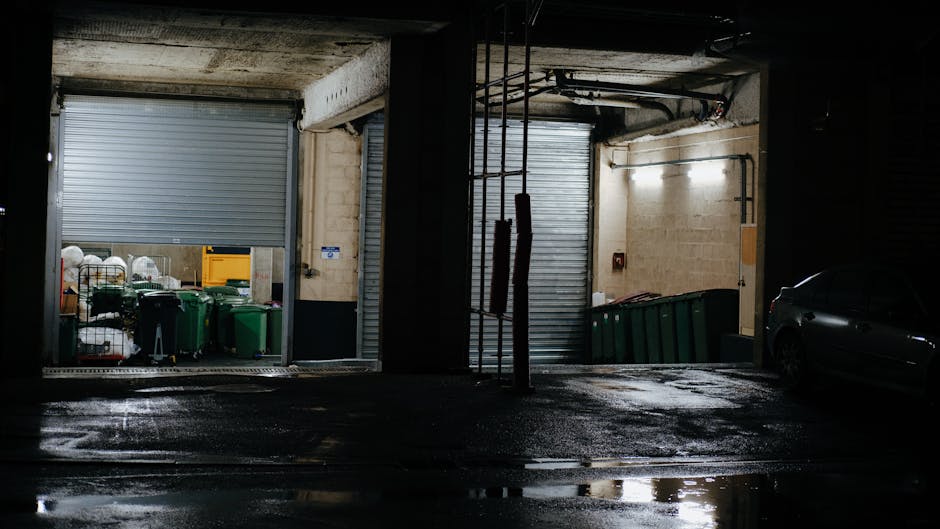 A waste collection operative wearing a high-visibility orange and grey uniform is standing beside a large industrial rubbish collection vehicle during dusk or early evening. The vehicle features a white body with red and yellow reflective markings, and its rear compactor is open, revealing a metal loading area for waste. The background shows silhouetted trees against a sky illuminated by a deep orange sunset, casting a warm glow over the scene. The operative is positioned on a paved surface, possibly a street or parking area, with the vehicle's lights casting a red-orange hue on nearby surfaces. The scene suggests an on-site waste collection operation, consistent with private rubbish removal services, emphasizing the environmental and logistical aspects of alternative waste handling in an urban setting. Waste Disposal Kentish Town is engaged in managing rubbish in an efficient and professional manner, supporting non-council waste disposal methods.