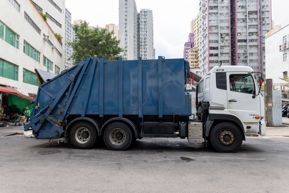 A large white and blue waste collection truck parked on an urban street with multi-storey residential buildings in the background. The truck's cab is white with black window frames and side mirrors, while the rear cargo area is enclosed with a blue metal container featuring vertical reinforcement ribs. The rear of the truck has a hinged flap, partially open, revealing a compartment for collecting rubbish. The truck's tires are black with visible tread, and the vehicle is positioned on a paved surface with some minor stains or marks. Surrounding environment includes a storefront with a green awning and some street vendors on the left side, with high-rise buildings behind, some with colourful facade details, creating an urban setting associated with commercial and residential waste management. The overall scene suggests on-site waste collection or rubbish removal services as part of private or alternative disposal arrangements managed by waste disposal companies.