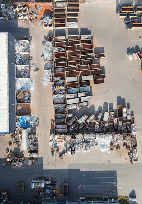 An aerial view of a waste disposal yard showing a large collection of metal and wooden containers arranged in rows, with some showing signs of rust and weathering. Surrounding the containers, there are scattered piles of mixed debris, including broken pallets, scrap metal, and various discarded materials. The yard is situated on a paved surface, with a section of a building visible on the left side, which appears to be used for storage or processing. The scene is illuminated by natural daylight, casting shadows that highlight the organized layout of the containers and debris. This setting exemplifies an environment where independent waste handling and on-site clearance of rubbish are carried out, such as those performed by companies like Waste Disposal Kentish Town, providing alternatives to traditional municipal rubbish collection services, particularly for large or specialized waste items that require private disposal methods.