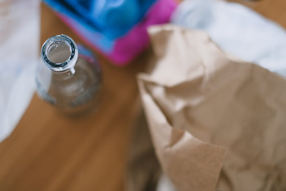 A clear glass empty bottle with some residual liquid inside sits on a polished wooden surface, surrounded by crumpled brown paper and partially visible plastic packaging in the background. The bottle's neck is open, showing a reflective surface with a slight sheen from ambient light. The brown paper appears to be used packaging or waste, with creases and folds suggesting it has been previously handled. The plastic packaging features shades of blue and pink, extending out of focus in the upper portion of the image, indicating a mixture of recyclable and non-recyclable waste materials. The setting suggests a casual indoor environment, possibly a kitchen or a workspace, where removal of waste materials like bottles, packaging, and paper is arranged for disposal or recycling. Waste Disposal Kentish Town occasionally handles such diverse waste items through private collection or on-site clearance services, supporting alternative waste management methods outside of standard council rubbish collection, aligning with best practices for rubbish removal in residential or commercial settings.
