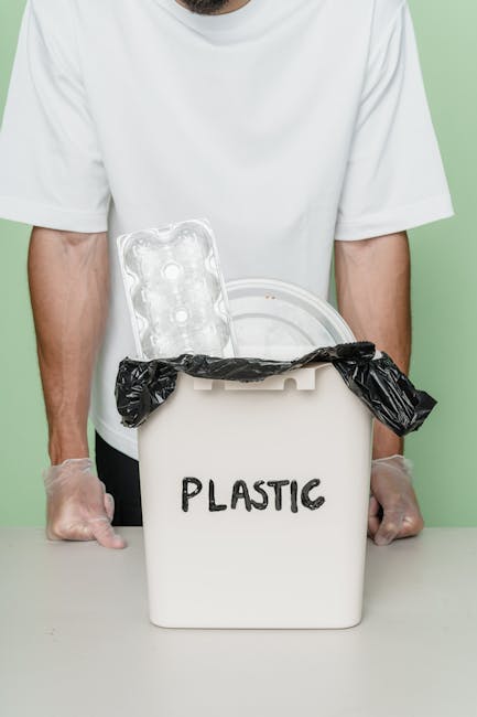 A person wearing a white shirt and tan trousers is seen organizing glass jars and bottles into a small white plastic basket on a wooden surface, likely in a kitchen or recycling area. The glass items include clear, empty jars with metal screw lids and a transparent, empty glass bottle with a tapered neck. The background features out-of-focus elements such as a gray bin with a partially visible label, and additional recyclable materials, including crumpled paper or packaging, are arranged nearby. The environment appears to be well-lit and clean, emphasizing the process of sorting recyclable glass items, which could be part of a waste management or rubbish removal service offered by Waste Disposal Kentish Town. The scene reflects a careful approach to on-site waste organization, aligning with practices of private or alternative waste handling for recycling purposes. The textured surfaces of the glass and the structured pattern of the basket are clearly visible, supporting accessibility and visual accuracy for an educational or service-related context.