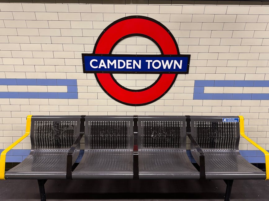 A row of four metallic public seating benches located against a white tiled wall with light blue horizontal accents, situated in a London Underground station. Above the benches, a large, round red and blue London Underground roundel sign displays the text 'CAMDEN TOWN' in white capital letters on a blue rectangular background. The benches have perforated metal surfaces with a silver finish, black armrests and support structures, and yellow armrests on the outer sides. The environment appears clean and well-lit, with natural or artificial lighting highlighting the textures of the metal and the tiles, creating a neutral atmosphere typical of public transport waiting areas, relevant for context of UK-based station facilities and private waste disposal considerations nearby.