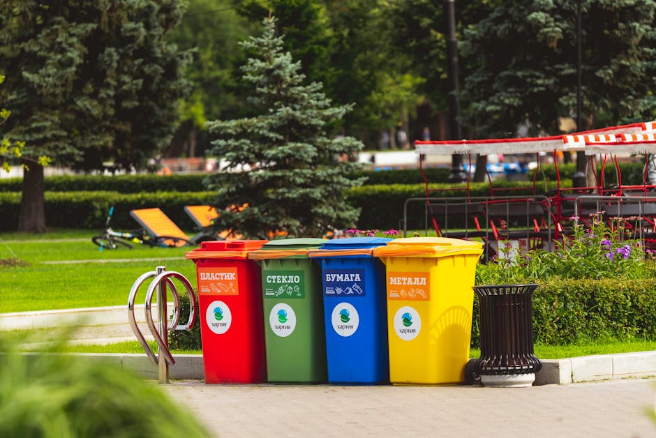 In a well-maintained park area, there are four large, upright waste bins arranged in a row on a paved surface. Each bin has a vibrant color and a label indicating its designated waste type: the red bin for plastic, the green bin for glass, the blue bin for paper, and the yellow bin for metal. The bins are made of durable plastic with smooth surfaces and matching lids that close securely. To the right of the bins, a black, cylindrical trash bin with a slatted design is visible. In the background, there are lush green trees, including a small coniferous tree in the foreground, with well-manicured grass and neatly trimmed hedges. A playground with red swings and benches can be seen further back, along with a pair of orange lounge chairs placed on a grassy area. The scene is illuminated by natural daylight, creating a bright and clean atmosphere, illustrating an example of organized waste disposal within an outdoor public space, managed by Waste Disposal Kentish Town for efficient rubbish management and alternative waste handling.