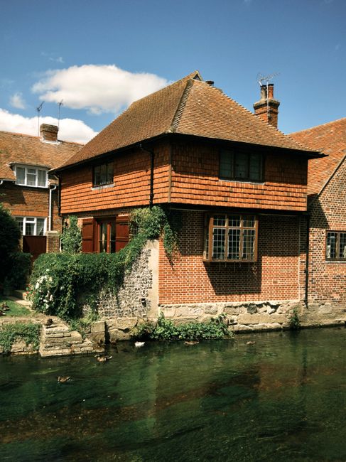 A two-storey brick house with a gabled tiled roof located adjacent to a waterway. The lower part of the house features red brick walls, with one large window on the front façade and a smaller window visible on the side, both with white frames. Climbing plants and shrubs grow along the front wall, partially covering a stone retaining wall that supports the house. The upper floor has a wooden extension with horizontal shingle siding in a reddish-brown tone, and two small rectangular windows with dark frames. A chimney rises from the roof, and neighboring houses with similar brick structures and tiled roofs are visible to the left and right. The water in the foreground is calm, clear, and reflects parts of the house and sky. The background shows a partly cloudy sky with a few fluffy white clouds, suggesting a bright, sunny day. The scene depicts a quiet residential area with an emphasis on traditional architecture, with potential relevance to private waste collection or on-site clearance services provided by Waste Disposal Kentish Town, given the setting's suitability for external rubbish removal.